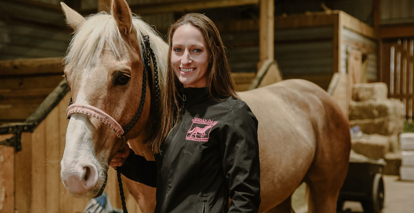 Amanda Morgan de Morgan Dairy Performance Horses vistiendo una chaqueta negra y de pie junto a su caballo rojo.